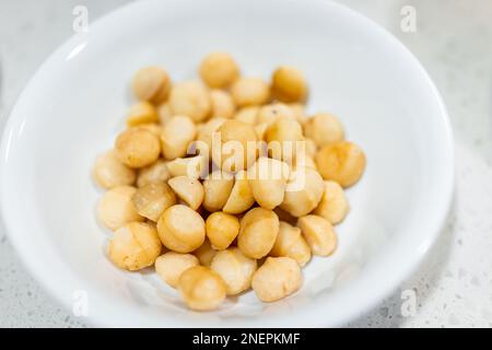 Macadamia-Nüsse geröstet und gesalzen, isoliert vor weißem Hintergrund, Schüssel auf dem Tisch mit Makroaufnahme der Konsistenz gelber Lebensmittel Stockfoto