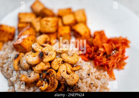 Gebackene Tofu-Würfel, gewürzte Cashews auf braunem Reis und eingelegter Kimchi-Kohl auf weißem Teller, mit strukturierten Details der Nahrung Stockfoto