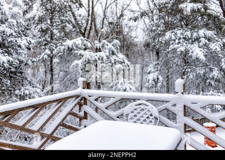 Weißer, schneebedeckter Tischstuhl aus Metall im Außenbereich auf der Holzterrasse des Hauses mit Geländerzaun im Winter mit Waldbäumen im Norden Virginias Stockfoto