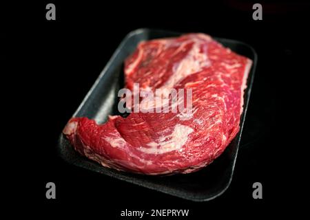 Verpacktes rohes rohes Rindfleisch, schwarz, angus, Tri-Tip-Steak-Nahaufnahme mit schwarzem Hintergrund, Studioaufnahme mit Fettmarmorierung und Tablett Stockfoto
