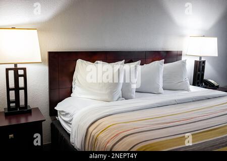 Schlafzimmerausstattung mit vier weißen Kissen mit Bettlaken auf Matratzenkopfteil im modernen Hotel mit leerem Zimmer und beleuchteten Lampen, Lampen und Nobod Stockfoto