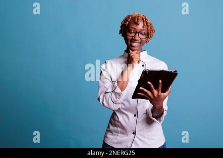 Afroamerikanische Küchenarbeiterin, die Rezepte-Videos auf dem Tablet-Bildschirm ansieht. Afro-Koch lächelt während der Arbeit Video-Unterricht auf elektronischem Gerät. Eine Frau kocht, die Spaß mit Technik hat. Stockfoto