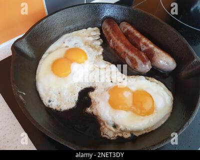 Doppelt gebratene Eier, sonnige Seite nach oben, gebratene Würstchen auf einer gusseisernen Bratpfanne auf einem Herd Stockfoto