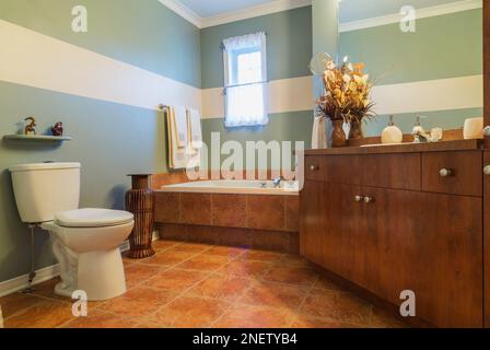 Rostfarbener Melaminholztisch mit geschichteter Arbeitsfläche und eingeschlossener Badewanne im oberen Badezimmer mit Keramikfußfliesen und großem Spiegel. Stockfoto