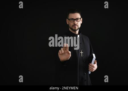 Priester mit Bibel macht Segen-Geste auf dunklem Hintergrund, Platz für Text Stockfoto