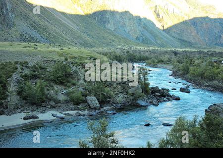 Das Bett eines wunderschönen Flusses fließt in einem stürmischen Bach, der sich entlang des Bodens eines tiefen Canyons im Schatten der Berge von der untergehenden Sonne zieht. Stockfoto