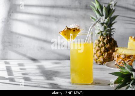 Ananassaft auf grauem Hintergrund. Es gibt Ananas- und Ananasscheiben in der Nähe, Sonnenlicht Stockfoto