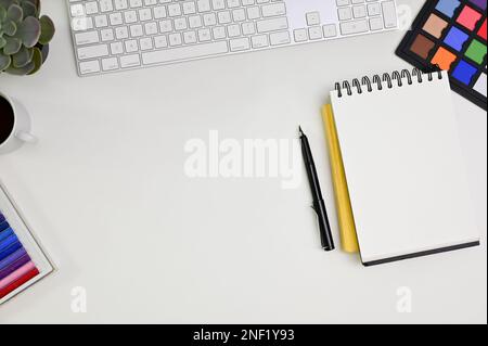 Moderner, weißer Tischhintergrund mit Tastatur, Farbprüfer, Schreibwaren, Stift, leerem Notizblock und Kopierbereich. Draufsicht, Grafikdesigner oder Stockfoto