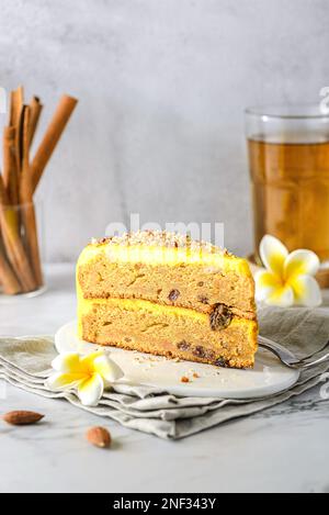 Ein Stück Karottenkuchen mit Tee. Der Kuchen liegt auf einem weißen Teller, verziert mit Mandeln, hinter einem Glas mit Zimtstangen und einem Glas Tee Stockfoto