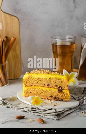 Ein Stück Karottenkuchen mit Tee. Der Kuchen liegt auf einem weißen Teller, verziert mit Mandeln, hinter einem Glas mit Zimtstangen und einem Glas Tee Stockfoto