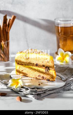 Ein Stück Karottenkuchen mit Tee. Der Kuchen liegt auf einem weißen Teller, verziert mit Mandeln, hinter einem Glas mit Zimtstangen und einem Glas Tee Stockfoto