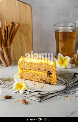 Ein Stück Karottenkuchen mit Tee. Der Kuchen liegt auf einem weißen Teller, verziert mit Mandeln, hinter einem Glas mit Zimtstangen und einem Glas Tee Stockfoto