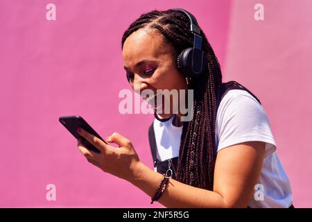 Ich habe noch nie etwas Lustigeres gesehen. Eine attraktive junge Frau, die an einer rosa Wand steht und lacht, während sie Technologie benutzt. Stockfoto