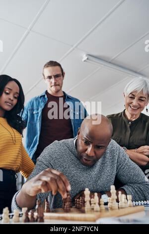 Er spielt immer, um zu gewinnen. Ein erwachsener Geschäftsmann, der in seinem Büro Schach spielt, während seine Kollegen im Hintergrund zuschauen. Stockfoto