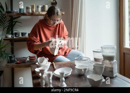 Fokussierte, konzentrierte Keramikarbeiterin, die in einem Handwerksgeschäft einen Tonartikel hält. Stockfoto