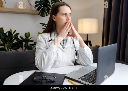 Beunruhigte junge Ärztin in weißer medizinischer Jacke und Stethoskop, die auf den seitlichen Laptop schaut. Unglücklicher Facharzt bei Spannungsgefühl und Müdigkeit Stockfoto