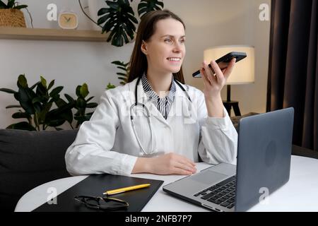 Lächelnde, schöne junge Ärztin, die Audio-Nachrichten auf einem Smartphone aufnimmt, am Tisch mit einem Laptop sitzt, mit dem Patienten online chattet, Beratung gibt Stockfoto