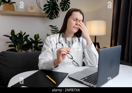 Aufgewühlte junge Ärztin in Uniform und Stethoskop Therapeutin am Tisch sitzender Arzt mit Augenbelastung, trockenes Auge Syndrom, Massage Stockfoto