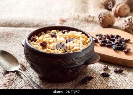 Süßer Hirse-Haferbrei mit dunklen Rosinen in rustikaler Keramikschüssel mit Walnüssen auf dem Hintergrund Stockfoto