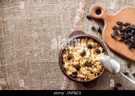 Süßer Hirse-Haferbrei mit dunklen Rosinen in rustikaler Keramikschüssel auf Sacktuchhintergrund mit Kopierbereich. Flach liegend, Draufsicht Stockfoto