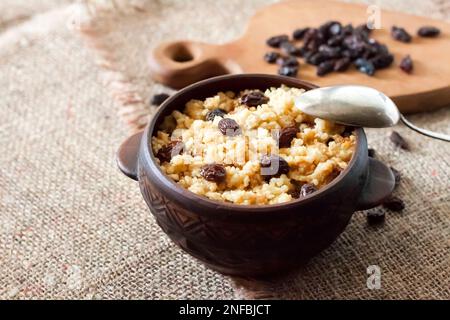 Süßer Hirse-Haferbrei mit dunklen Rosinen in rustikaler Keramikschüssel mit Walnüssen auf dem Hintergrund Stockfoto