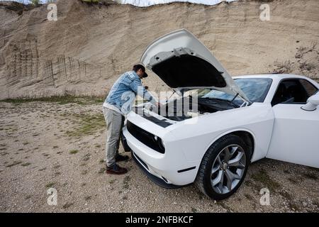 Ein hübscher Mann in Jeans-Jacke und -Mütze steht neben seinem weißen Muskelauto mit sich öffnender Kapuze, Ölstand des Motors überprüfen. Stockfoto