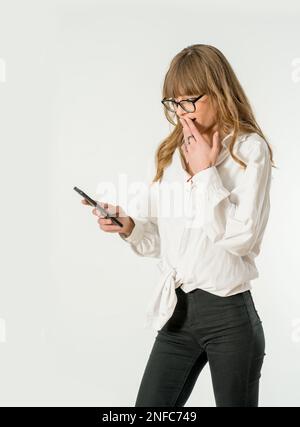 Eine junge Frau mit Brille steht vor weißem Hintergrund, ihr Blick konzentrierte sich auf ihr Handy Stockfoto