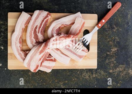 Nahaufnahme von vier Streifen ungekochten Speck, der auf einem Holzbrett brutzelt Stockfoto