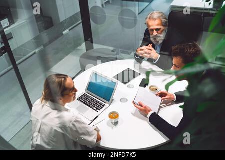 Blick von oben auf drei Geschäftsleute, die am runden Tisch sitzen und ein Geschäftstreffen in einem gläsernen Büro abhalten Stockfoto