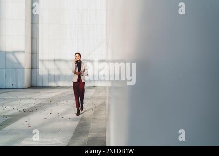 Junge Geschäftsfrau, die an einem sonnigen Tag an ihrem Bürogebäude vorbeigeht und telefoniert Stockfoto