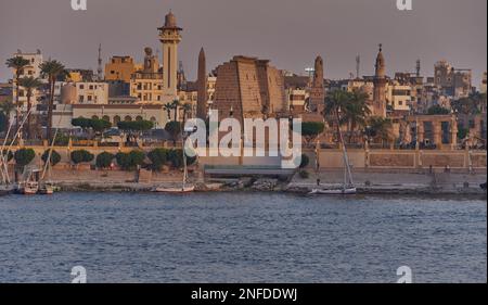 Luxor, ägyptischer Sonnenuntergang vom Westufer aus, der den Nil mit Felukken, Kreuzfahrtschiffen und dem Luxor Tempel am Ostufer zeigt Stockfoto