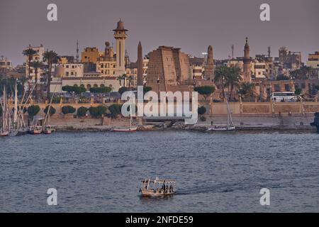 Luxor, ägyptischer Sonnenuntergang vom Westufer aus, der den Nil mit Felukken, Kreuzfahrtschiffen und dem Luxor Tempel am Ostufer zeigt Stockfoto