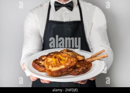 Ein Kellner in Schürze und Fliege zeigt einen Teller Gourmet-Torrijas. Ein typisch spanischer Süßstoff mit Brot, Milch, Eiern, Zucker und Zimt Stockfoto