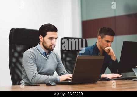 Ein Geschäftsmann, der am Schreibtisch sitzt und an seinem Laptop und Computer arbeitet, mit einem fokussierten Gesichtsausdruck Stockfoto
