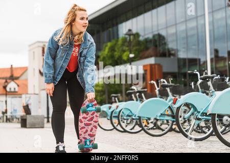 Lächelndes, blondes Mädchen mit einer Jeans in der Hand, das ein Skateboard mit lustigem Aufdruck darunter hält und sich auf das Skaten in der Stadt vorbereitet Stockfoto