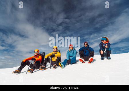 Gruppe von fünf Bergsteigern, die hoch oben in den verschneiten Alpen in der Sonne sitzen Stockfoto