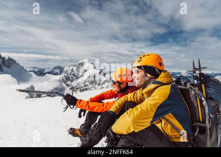 Seitlicher Blick auf zwei lächelnde Wanderer in Winterausrüstung, die an einem sonnigen Tag hoch oben in den Bergen im Schnee sitzen Stockfoto