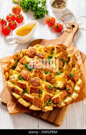 Käsiges Knoblauchbrot, das ganze runde Brot auf dem Schneidebrett auf dem weißen Holztisch auseinanderziehen, mit geschmolzener Butter, Tomaten und Gewürzen im Hintergrund, vertica Stockfoto