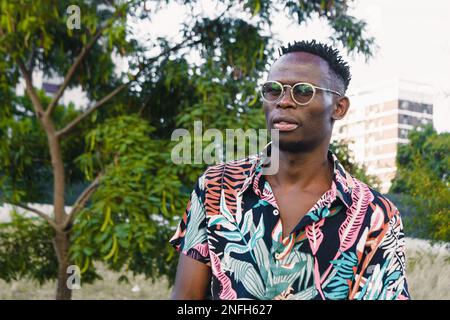 Porträt eines jungen Mannes afrikanischer ethnischer Herkunft, der Brille und farbenfrohe Kleidung trägt, auf einem öffentlichen Platz steht und draußen in die Kamera schaut, mit Tre Stockfoto