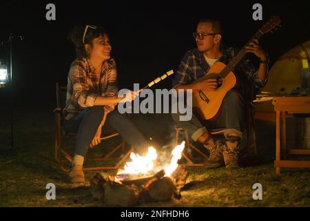 Asiatische Frau, die Marshmallows anstoßt, während ihr Freund am Lagerfeuer Gitarre spielt, wo sie ihre Zelte für das Camping am See nachts aufstellen. Stockfoto