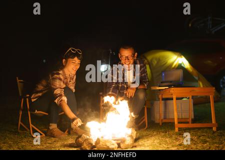 Asiatisches Paar grillt Mais an einem Feuer, das sie für die Nacht eingerichtet haben, während sie mitten in einem Zelt in der Nähe eines Sees campen. Stockfoto