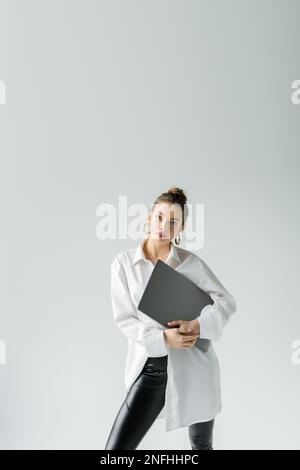 Trendige Frau in weißem Hemd und Ring-Ohrringen posiert mit Laptop und schaut isoliert auf die Kamera, auf grauem Stockbild Stockfoto