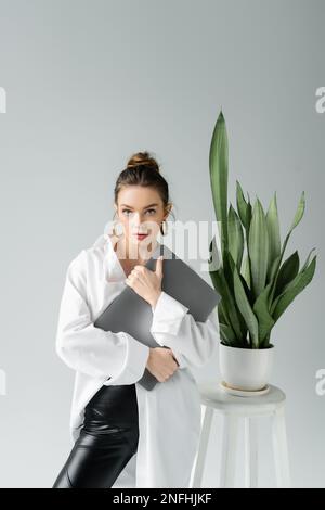 Trendige Frau mit Laptop, die in der Nähe eines Blumentopfes auf die Kamera schaut, mit tropischer Pflanze isoliert auf grauem Stockbild Stockfoto