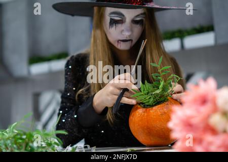 Ein Mädchen in einem Hexenkostüm und mit einem bemalten Gesicht kreiert eine Blütenzusammensetzung aus einem Kürbis. Hochwertiges Foto Stockfoto