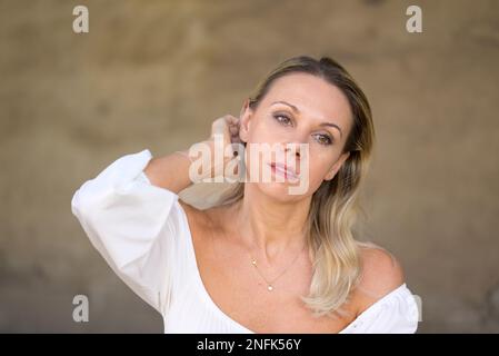 Das Porträt einer blonden Frau mittleren Alters mit einer weißen Bluse führt eine Hand durch ihr Haar mit einem durchdachten Gesichtsausdruck vor einer braunen Wand Stockfoto