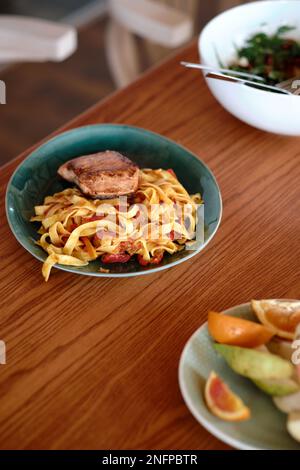 Ein hausgemachtes Abendessen mit Thunfisch mit scharfen flachen Nudeln, Salat und Obst auf einem Holztisch mit weißen Stühlen im Hintergrund Stockfoto