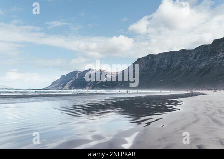 Reflexionen in feuchten Sand und Playa de Famara Strand auf Lanzarote, Kanarische Inseln, gegen Berge, Meer und blauem Himmel Stockfoto