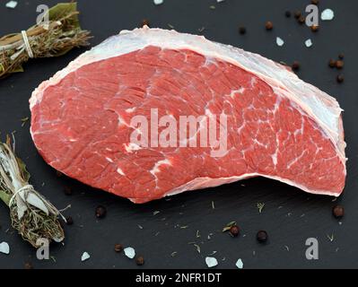 Frisches rohes Rumpffleisch, das von der Picanha-Kappe auf einer schwarzen Schieferplatte geschnitten wurde, mit Salz, Pfeffer und Gewürzbündel Stockfoto