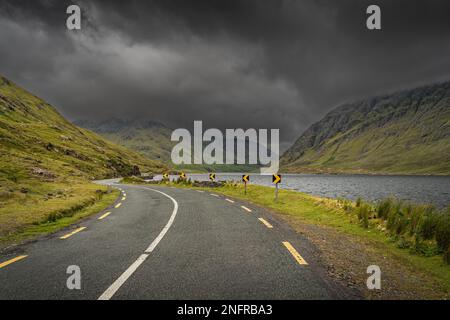 Gewundene Straße führt durch das Doolough Valley mit See, zwischen Glenummera und Glencullin Gebirgsketten mit dunklem dramatischen Himmel, Mayo, Irland Stockfoto