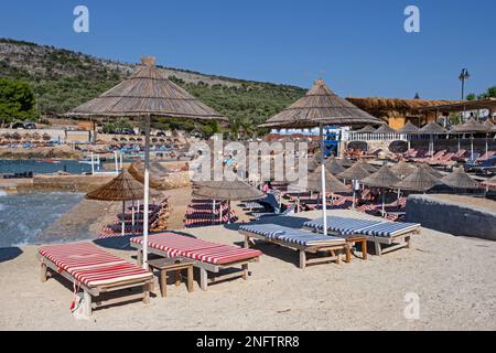 Sonnenschirme und leere Sonnenliegen am Sandstrand des Badeorts Ksamil entlang der albanischen Riviera im Sommer, Sarandë, Kreis Vlorë, Südalbanien Stockfoto